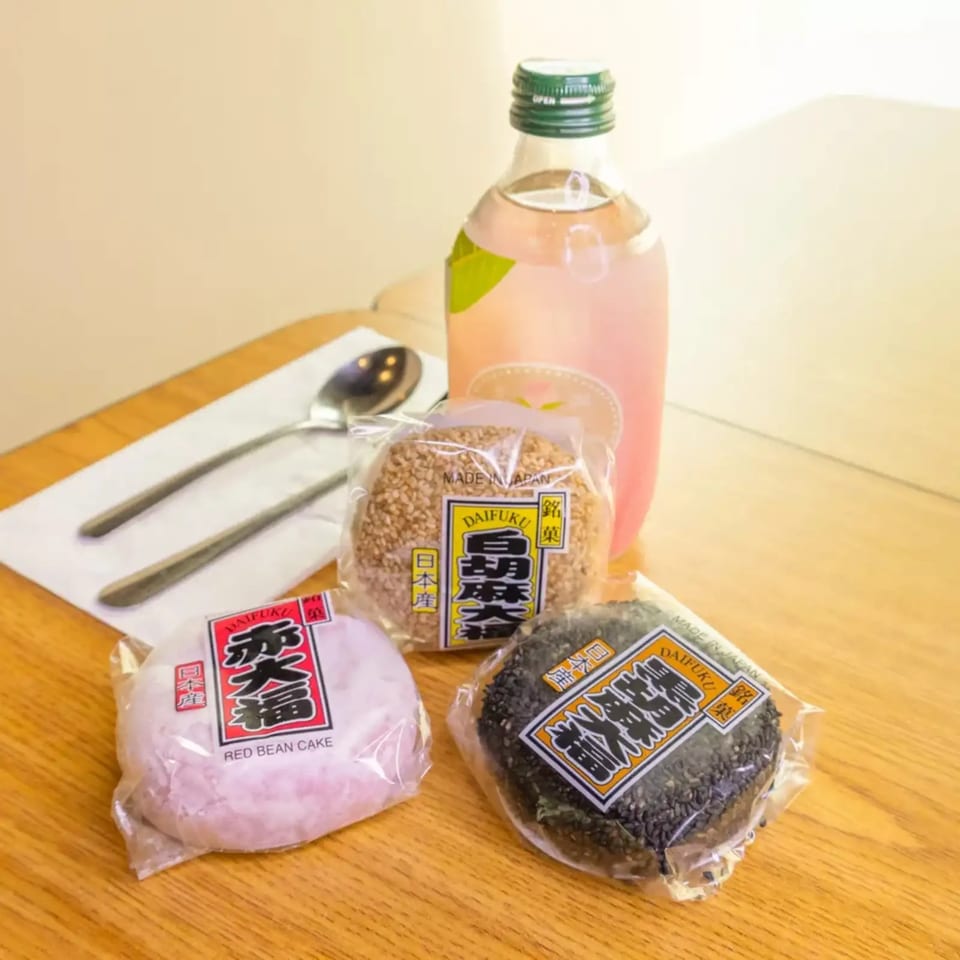 Three packaged Daifuku mochi, red bean, sesame, and black sesame, with a pink drink on a wooden table.