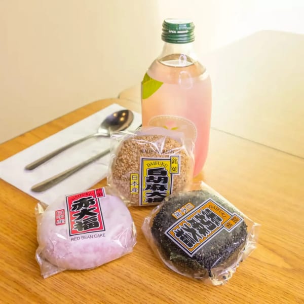 Three packaged Daifuku mochi, red bean, sesame, and black sesame, with a pink drink on a wooden table.