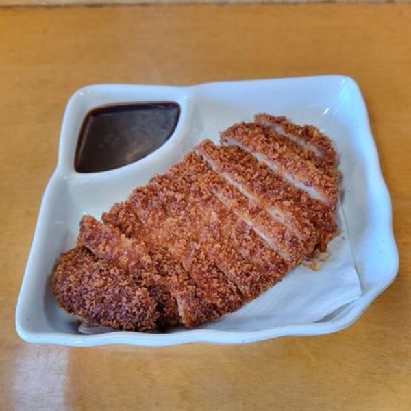 Crispy, sliced tonkatsu with a side of dark dipping sauce on a white rectangular plate.