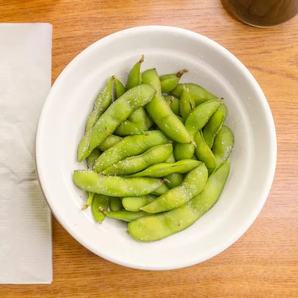 A white bowl filled with bright green edamame pods, lightly sprinkled with sea salt, on a wooden table.