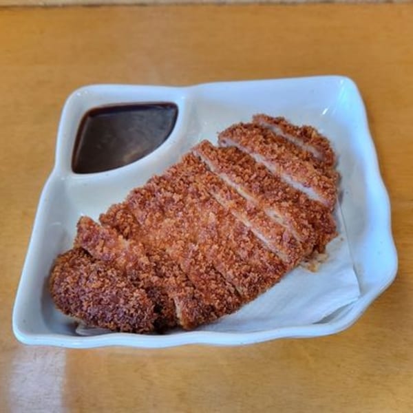 Crispy, sliced tonkatsu with a side of dark dipping sauce on a white rectangular plate.