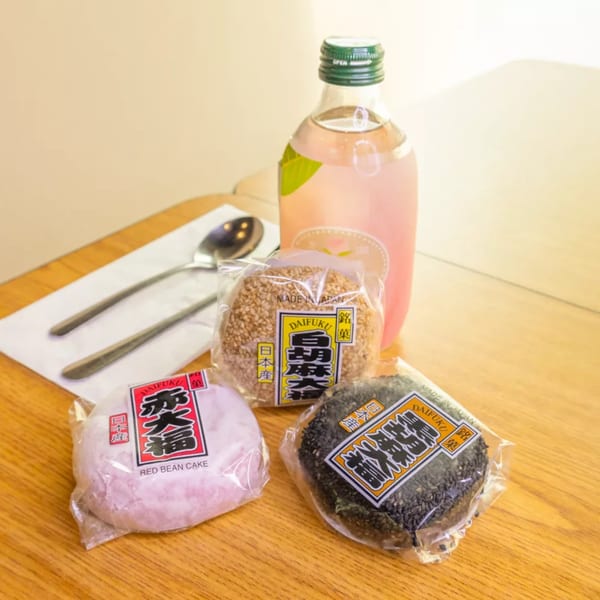 Three packaged Daifuku mochi, red bean, sesame, and black sesame, with a pink drink on a wooden table.