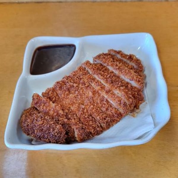 Crispy, breaded pork cutlet (tonkatsu) slices with a side of dark dipping sauce on a white plate.